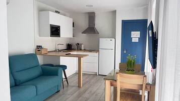 Une cuisine compacte avec des meubles blancs, un four à micro-ondes et une cuisinière. On y trouve une porte bleue et une petite table avec une chaise.