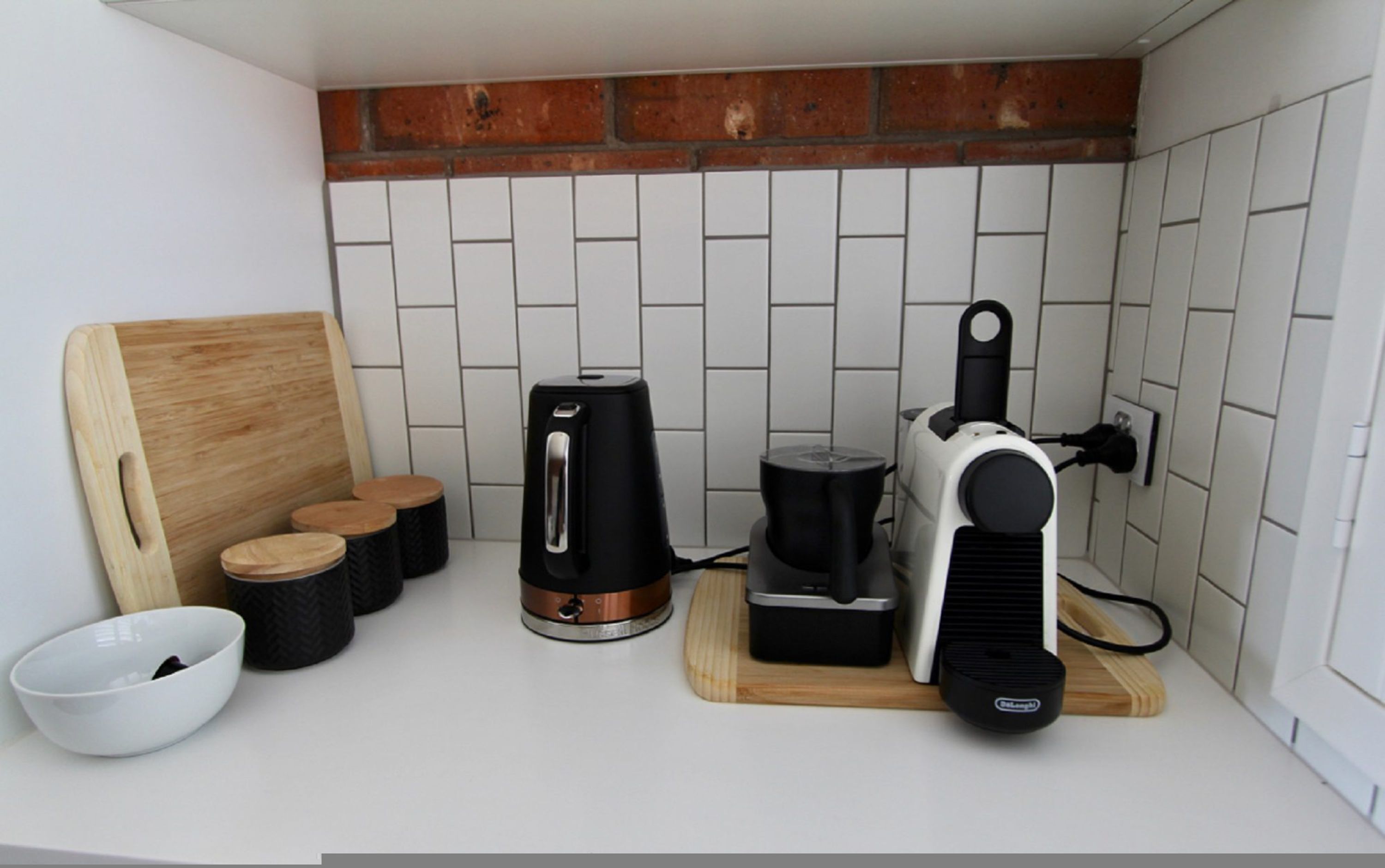 A kitchen with a brick backsplash, a wooden cutting board, a coffee maker, a kettle, and a rice cooker.