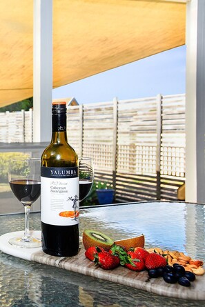 Una bottiglia di Yalumba Cabernet Sauvignon, due bicchieri di vino rosso e un vassoio di frutta su un tavolo con vista su una recinzione di legno.
