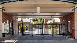 Une entrée fermée par un portail mène à un bâtiment doté d’une allée couverte, de murs en briques et offrant une vue dégagée sur les voitures garées et la verdure en arrière-plan.