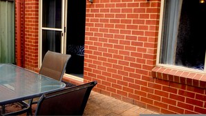 Une table en verre avec des chaises, placée à l’extérieur d’un bâtiment en briques, une porte-fenêtre coulissante et une fenêtre donnant sur le ciel.