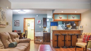 A living room with a sofa, a red ottoman, a kitchen area with bar stools, and a dining table. - The Red Chair Retreat at Red River (Red River)