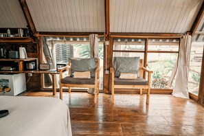 Une piĂšce confortable avec un lit, deux chaises en bois avec des coussins, une petite kitchenette et une grande fenĂȘtre avec des rideaux.