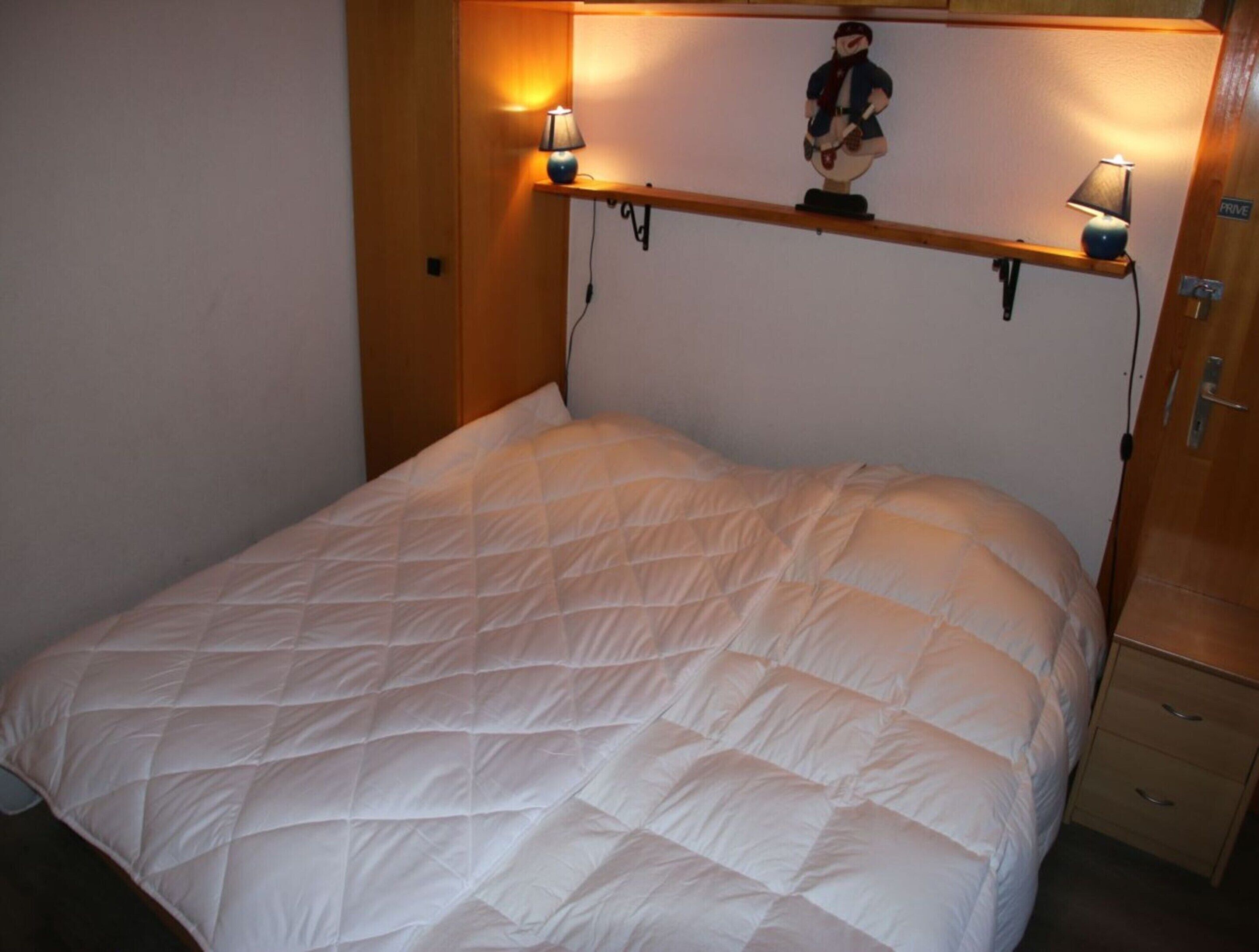 A neatly made bed with a white quilted duvet, two bedside lamps, and a small shelf with a figurine.