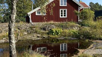 A red wooden house with two windows and a flag, situated by a calm body of water with surrounding vegetation.