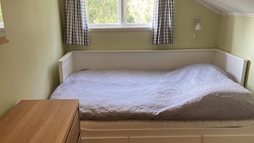 A single bed with a white headboard and drawers, a wooden nightstand, a window with checkered curtains, and a wall-mounted lamp.