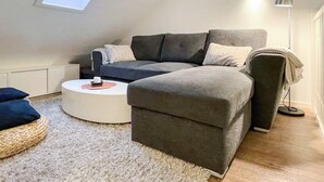 Un moderno loft con un ampio divano angolare grigio, un tavolino da caffè bianco rotondo e un piccolo tavolino laterale. Un lucernario offre luce naturale e una lampada da terra si trova a destra.