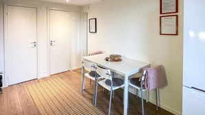 Una cucina compatta con un tavolo bianco, due sedie e un frigorifero. Si notano due porte e un piccolo quadro appeso al muro.