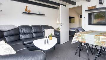 A modern living room with a black leather sofa, a white coffee table, and a dining table with a patterned tablecloth. There is a television mounted on the wall and a door leading to another room.