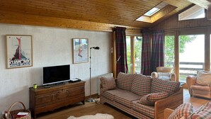 A cozy living room with wooden ceiling beams, a plaid sofa, a wooden cabinet with a flat-screen TV, and a large window with curtains. - Chalet With Mesmerizing Mountain View (Veysonnaz)