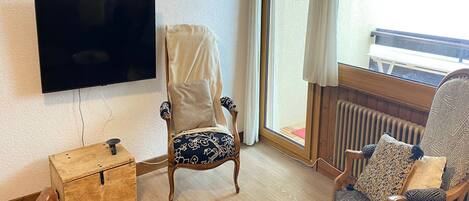 A living room with a flat-screen TV mounted on the wall, two wooden chairs with patterned upholstery, a small round table with a candle, and a wooden chest of drawers.