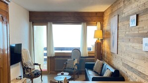 A cozy living room with a wooden wall, a sofa, a coffee table, a TV, and a view of the outdoors. - Ski Lift Apartment With Views (Veysonnaz)