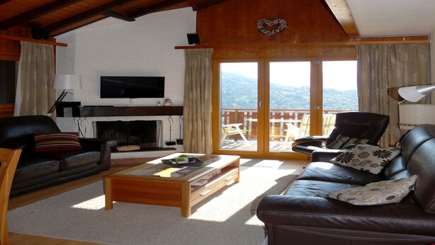 A cozy living room with a fireplace, leather sofas, a coffee table, and a large window offering a mountain view. - Apartment for Relaxed Vacation (Veysonnaz)