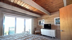 A bedroom with a bed, a dresser, a television, and a chair. There is a sliding glass door leading to an outdoor area with a chair. - Charming 2 bedroom direct oceanfront townhouse in family friendly midtown. (Ocean City)