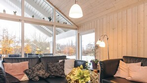 A cozy living room with a black leather sofa, patterned cushions, a vase with flowers, and large windows offering a view of trees and a snowy landscape.
