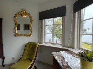 Une pièce comprenant un fauteuil recouvert d’un tissu vert, un bureau en bois surmonté d’une nappe en dentelle, un miroir à cadre doré et une fenêtre donnant sur un paysage verdoyant.