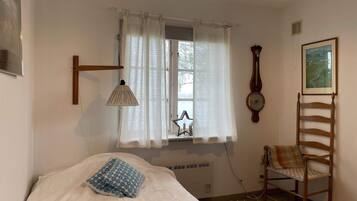 Un dormitorio con cama, una silla, una lámpara, una ventana con cortinas, un reloj y un cuadro en la pared.