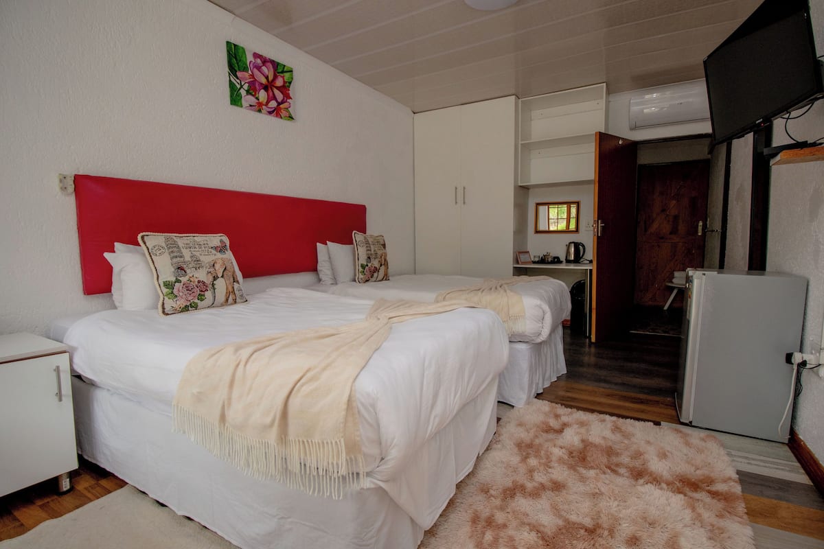A bedroom with two beds, a red headboard, a white wardrobe, a small kitchenette, and a television.
