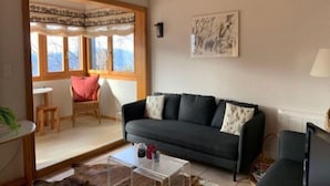Ein Wohnzimmer mit einem dunkelgrauen Sofa, einem transparenten Glastisch, Holzboden und einem Fenster mit Blick auf Bäume.