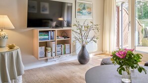 Ett modernt vardagsrum med en platt-TV, bokhyllor, en vas med blommor och utsikt över naturen.