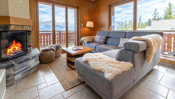 A cozy living room with a stone fireplace, a grey sofa, and a view of mountains through large windows.