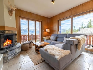 A cozy living room with a stone fireplace, a grey sofa, and a view of mountains through large windows. - Hauts de Veysonnaz HV2 010 -sauna Apartment 6 Pers (Veysonnaz)