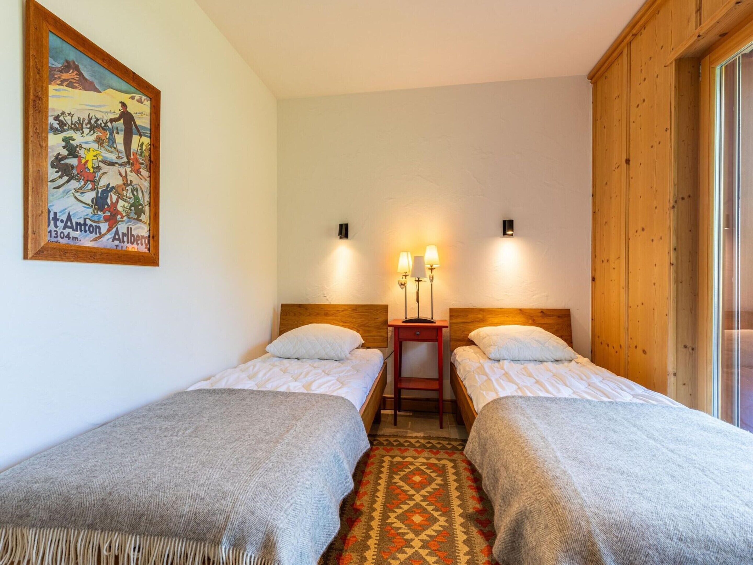 A hotel room with two single beds, a wooden headboard, a small bedside table with lamps, a patterned rug, and a painting on the wall.