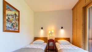 A hotel room with two single beds, a wooden headboard, a small bedside table with lamps, a patterned rug, and a painting on the wall.