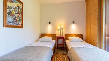 A hotel room with two single beds, a wooden headboard, a small bedside table with lamps, a patterned rug, and a painting on the wall.