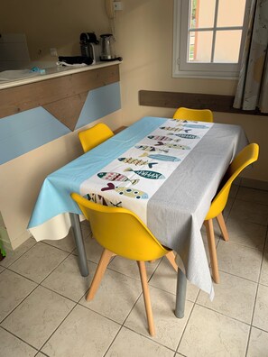 Un coin repas avec une table recouverte d’une nappe ornée de motifs de poissons, entourée de chaises jaunes aux pieds en bois.