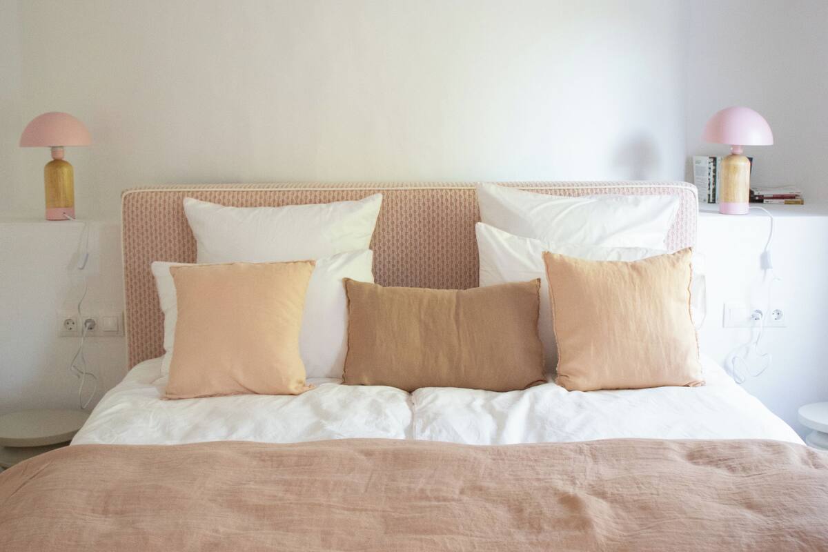 A neatly made bed with a pink and white headboard, accompanied by bedside lamps and a small shelf with books.