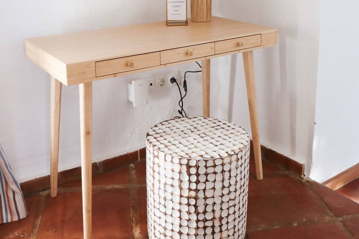A wooden desk with a lamp and a framed certificate, a round stool, and a power outlet on a tiled floor.