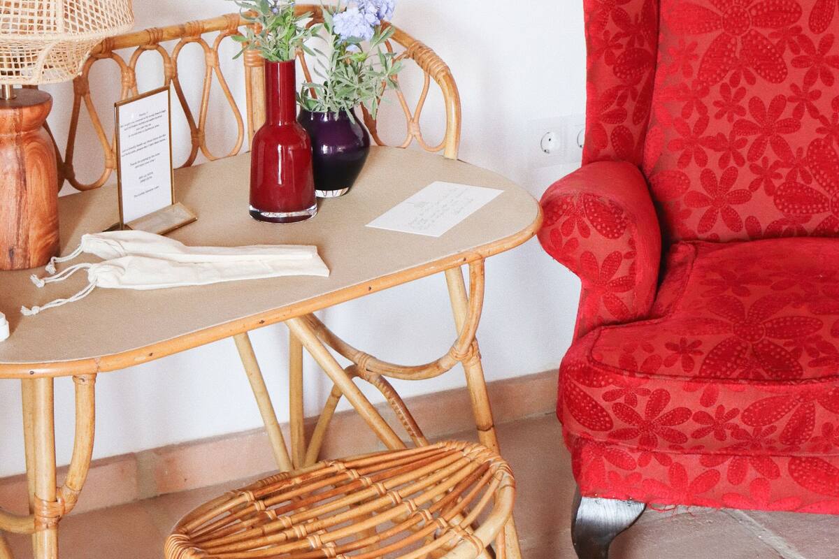 A room with a wicker table, two chairs, a red upholstered armchair, and two vases with flowers.