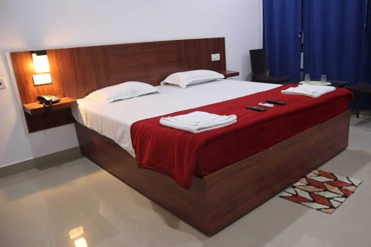 Una cama bien hecha con una colcha roja, almohadas blancas y un cabecero de madera. Se ve una mesita de noche con una lámpara y un teléfono, una pequeña mesa con una silla, y una cortina azul al fondo.