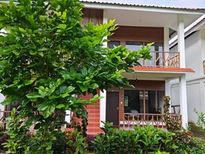 Ein zweigeschossiges Gebäude mit Balkon, Holzzaun und verschiedenen Pflanzen im Vordergrund.
