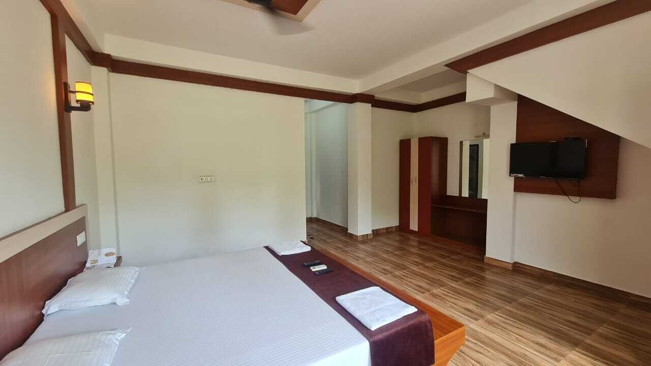 A hotel room with a large bed, a wooden headboard, a bedside table, a flat-screen TV mounted on the wall, and a wooden floor.