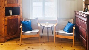 Ein gemütliches Zimmer mit einem Holzschrank, zwei Stühlen mit blauen Kissen, einem kleinen Tisch und einem Fenster mit Blick auf Bäume.