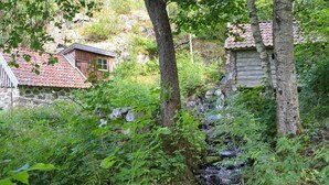 Ein rustikales Blockhaus mit Steinaußenwand und rotem Ziegeldach, umgeben von üppigem Grün und einem kleinen Bach.