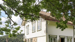 Ein zweistöckiges Haus mit Steinuntergrund, weißer Fassade und einem Ziegeldach. Vor der Terrasse befindet sich Gartenmöbel und im Hintergrund ist ein See mit bewaldeten Hügeln zu sehen.