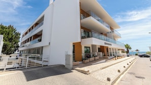 Un immeuble moderne à plusieurs étages, doté de balcons, d’un espace d’entrée pavé et offrant une vue dégagée sur la mer.