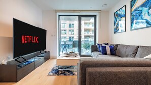 A modern living room with a flat-screen TV displaying 'NETFLIX,' a sofa, a coffee table, and a view of an apartment building through a glass door. - LUX Riverside Apartment in Royal Wharf I Near LCY Airport/Canary Wharf/ExCel/O2 (Greater London)