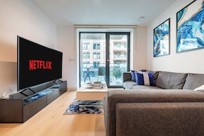 Una sala de estar moderna con un televisor de pantalla plana mostrando 'NETFLIX', un sofá, una mesa de centro y vista a un edificio de apartamentos por una puerta de vidrio.
