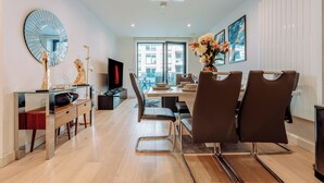A modern dining area with a glass table, brown chairs, a large mirror, and a vase of flowers. - LUX Riverside Apartment in Royal Wharf I Near LCY Airport/Canary Wharf/ExCel/O2 (Greater London)
