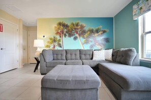 A living room with a grey sectional sofa, a matching ottoman, a lamp, and a wall mural of palm trees.