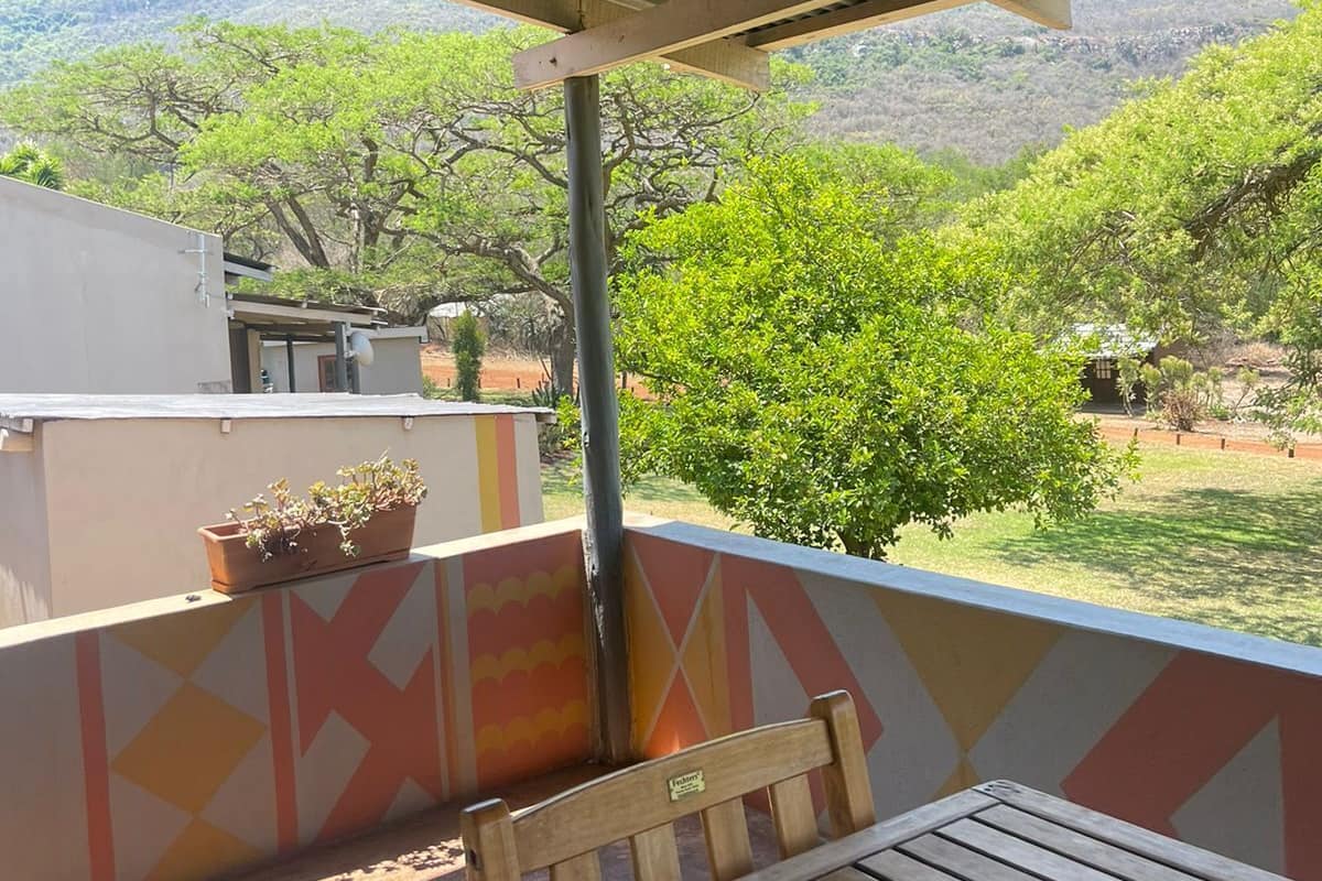 Un espacio al aire libre techado con un banco de madera y una mesa, plantas en macetas, y vistas a árboles y colinas verdes.