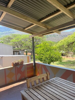 A covered outdoor area with a wooden bench and a table, potted plants, and a view of green trees and hills. - Madi a Thavha Mountain Lodge (Louis Trichardt)