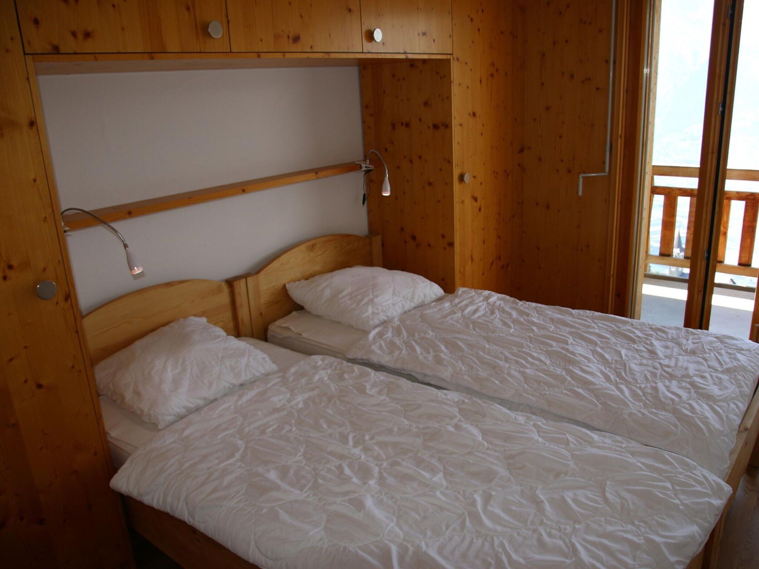 A wooden cabin room with two single beds, a shelf above the beds, and a window with a view of the outside.