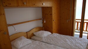Quarto de cabana de madeira com duas camas individuais, uma prateleira sobre as camas e uma janela com vista para o exterior.