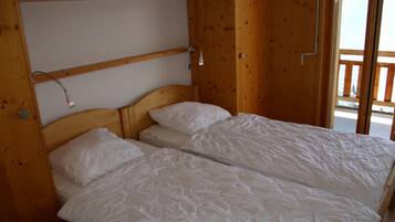 A wooden cabin room with two single beds, a shelf above the beds, and a window with a view of the outside.