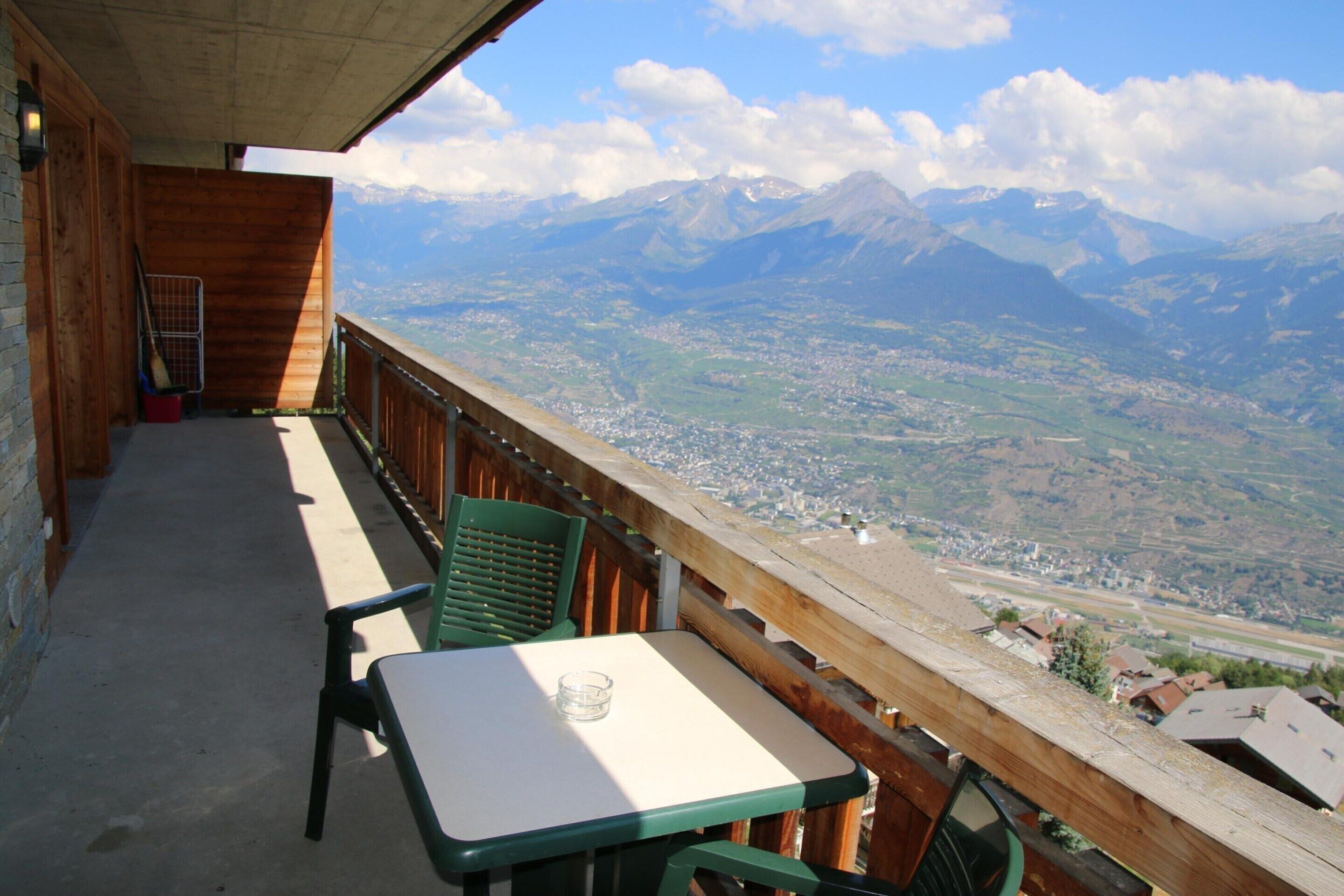 A balcony with a table and chairs, overlooking a mountainous landscape with a town below.